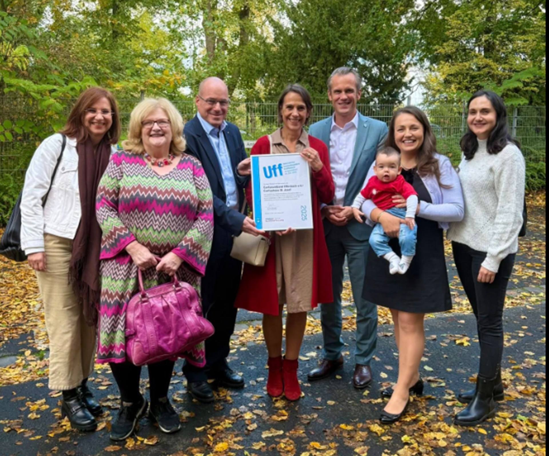 Gruppenbild, das Caritashaus-Mitarbeitende zusammen mit Caritasdirektor Martin Webers und dem Oberbürgermeister Felix Schwenke in einer herbstlichen Grünanlage zeigt. In der Mitte steht Bereichsleiterin Anette Bacher, die eine Urkunde hochhält. Eine andere Mitarbeiterin hält ein Baby im Arm. 