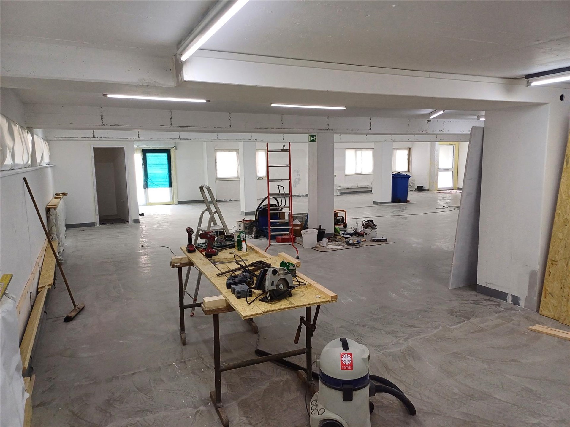 Renovierungsbereich in einem großen Raum mit grauem Boden und weißen Wänden. In der Mitte steht ein Arbeitstisch mit Werkzeugen wie einer Kreissäge. Im Vordergrund ein Staubsauger, im Hintergrund eine rote Leiter und weitere Baumaterialien.